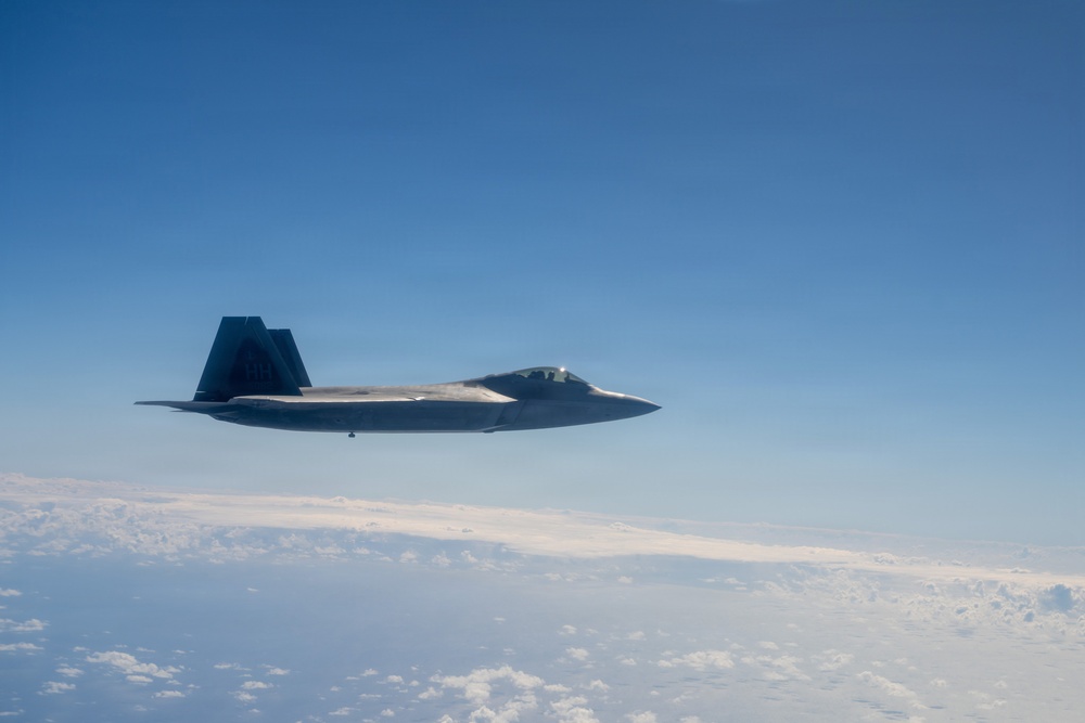 A single stealth fighter jet, consistent with the design of an F-22 Raptor, is photographed in level flight at high altitude above a cloud layer. The aircraft is viewed from a slightly elevated lateral angle, likely from another aircraft flying in formation. The sky is clear blue above with a scattered cloud deck visible below.