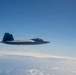 A single stealth fighter jet, consistent with the design of an F-22 Raptor, is photographed in level flight at high altitude above a cloud layer. The aircraft is viewed from a slightly elevated lateral angle, likely from another aircraft flying in formation. The sky is clear blue above with a scattered cloud deck visible below.
