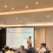 A presenter is delivering a training session in a hotel conference room, standing before a projected slide titled 'The Basics of Delivering a Brief.' The slide outlines the ABC framework: Accuracy, Brevity, and Clarity, with a logo suggesting a military or defense training organization. Several attendees are seated at tables with laptops, water bottles, and notepads.
