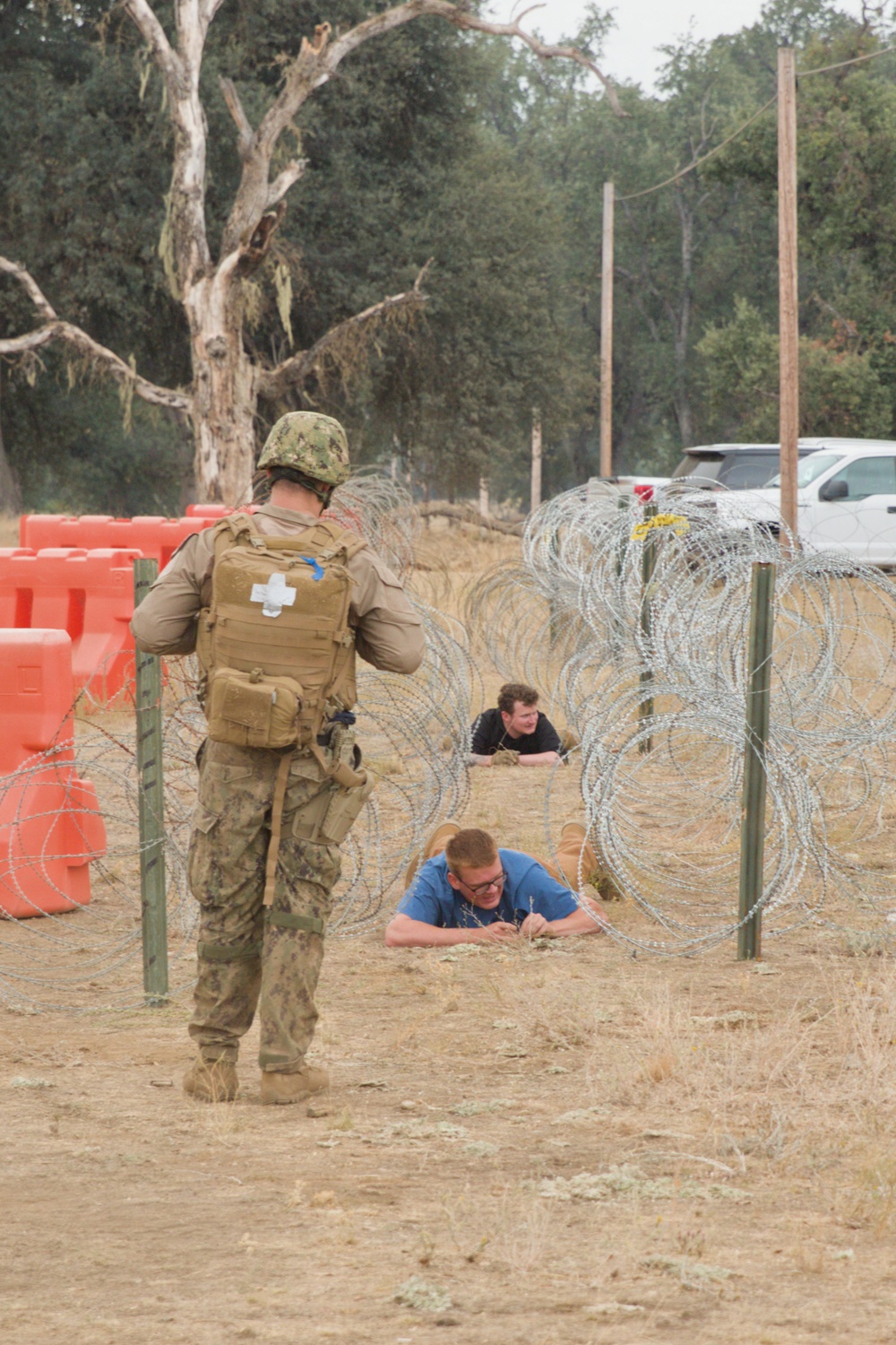 A military training exercise is underway in an outdoor field setting. A uniformed service member wearing a medical cross on their backpack supervises two individuals in civilian clothing crawling under concertina wire obstacles. The scene appears to be a controlled obstacle course training environment.