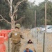 A military training exercise is underway in an outdoor field setting. A uniformed service member wearing a medical cross on their backpack supervises two individuals in civilian clothing crawling under concertina wire obstacles. The scene appears to be a controlled obstacle course training environment.