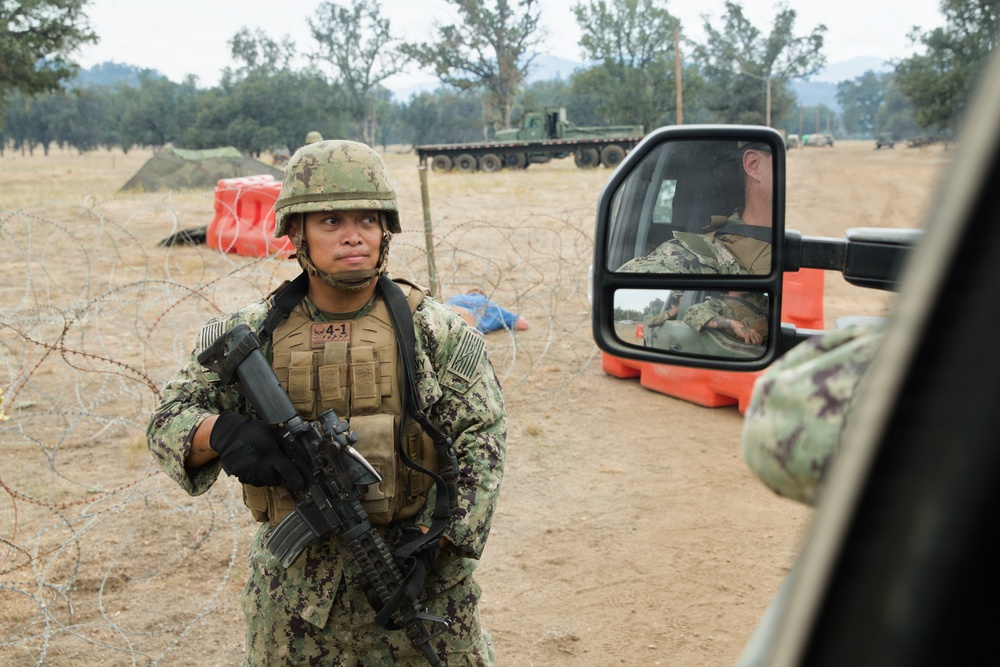 A U.S. Navy service member in NWU Type III (woodland digital camouflage) uniform stands at what appears to be a military checkpoint or training exercise area, holding an M4-style carbine rifle. The scene includes concertina wire barriers, orange barricades, and military vehicles in the background. A vehicle side mirror reflects another service member seated inside a truck.