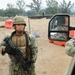A U.S. Navy service member in NWU Type III (woodland digital camouflage) uniform stands at what appears to be a military checkpoint or training exercise area, holding an M4-style carbine rifle. The scene includes concertina wire barriers, orange barricades, and military vehicles in the background. A vehicle side mirror reflects another service member seated inside a truck.