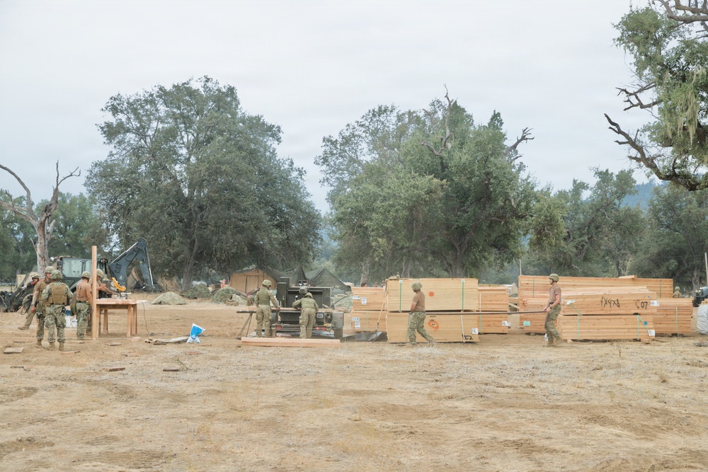 Military personnel in camouflage uniforms are engaged in construction or engineering activities at an outdoor field site. Large stacks of lumber (visible markings suggest 4x4 dimensions) are being handled and organized, with heavy equipment including what appears to be a backhoe visible in the background. The setting appears to be a dry, open area surrounded by oak trees, consistent with a military training or field operations environment.