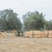 Military personnel in camouflage uniforms are engaged in construction or engineering activities at an outdoor field site. Large stacks of lumber (visible markings suggest 4x4 dimensions) are being handled and organized, with heavy equipment including what appears to be a backhoe visible in the background. The setting appears to be a dry, open area surrounded by oak trees, consistent with a military training or field operations environment.