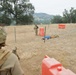 A military training exercise is taking place in an open field with dry grass and oak trees. Personnel in camouflage uniforms are positioned around a concertina wire perimeter marked with orange barriers, with a civilian role-player lying on the ground near the wire. The scene depicts a simulated crowd control or border security scenario.