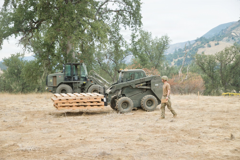 A military personnel in camouflage uniform and helmet walks alongside two olive-drab military construction vehicles in an open dry field. A CAT skid steer loader is carrying a stack of wooden pallets, with a larger wheeled front loader positioned behind it. The setting appears to be a rural training or operational area with oak trees and hills visible in the background.