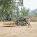 A military personnel in camouflage uniform and helmet walks alongside two olive-drab military construction vehicles in an open dry field. A CAT skid steer loader is carrying a stack of wooden pallets, with a larger wheeled front loader positioned behind it. The setting appears to be a rural training or operational area with oak trees and hills visible in the background.