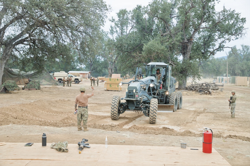 Military personnel are conducting construction or engineering operations at what appears to be a forward operating base or field camp. A motor grader is being directed by a service member while others observe or stand guard in the background. The scene includes various military equipment, supplies, and temporary structures set among large oak trees.