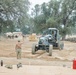 Military personnel are conducting construction or engineering operations at what appears to be a forward operating base or field camp. A motor grader is being directed by a service member while others observe or stand guard in the background. The scene includes various military equipment, supplies, and temporary structures set among large oak trees.