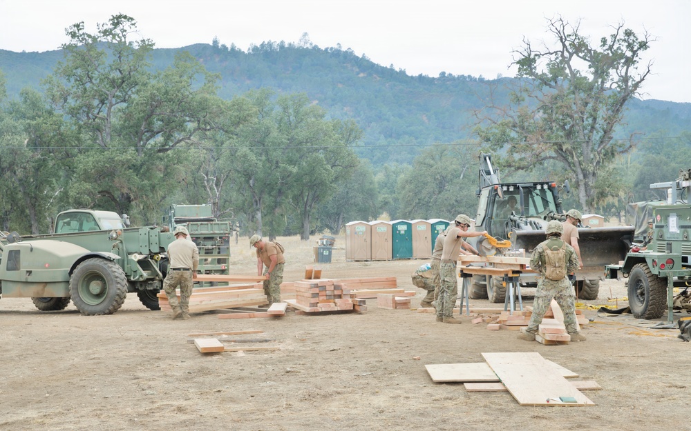 Military personnel in camouflage uniforms are engaged in construction or carpentry work at an outdoor field site, cutting and organizing lumber. The scene includes multiple military vehicles, heavy equipment, and portable toilets, suggesting a temporary forward operating base or training exercise. The setting is a dry, open area surrounded by oak trees and hills.