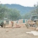 Military personnel in camouflage uniforms are engaged in construction or carpentry work at an outdoor field site, cutting and organizing lumber. The scene includes multiple military vehicles, heavy equipment, and portable toilets, suggesting a temporary forward operating base or training exercise. The setting is a dry, open area surrounded by oak trees and hills.