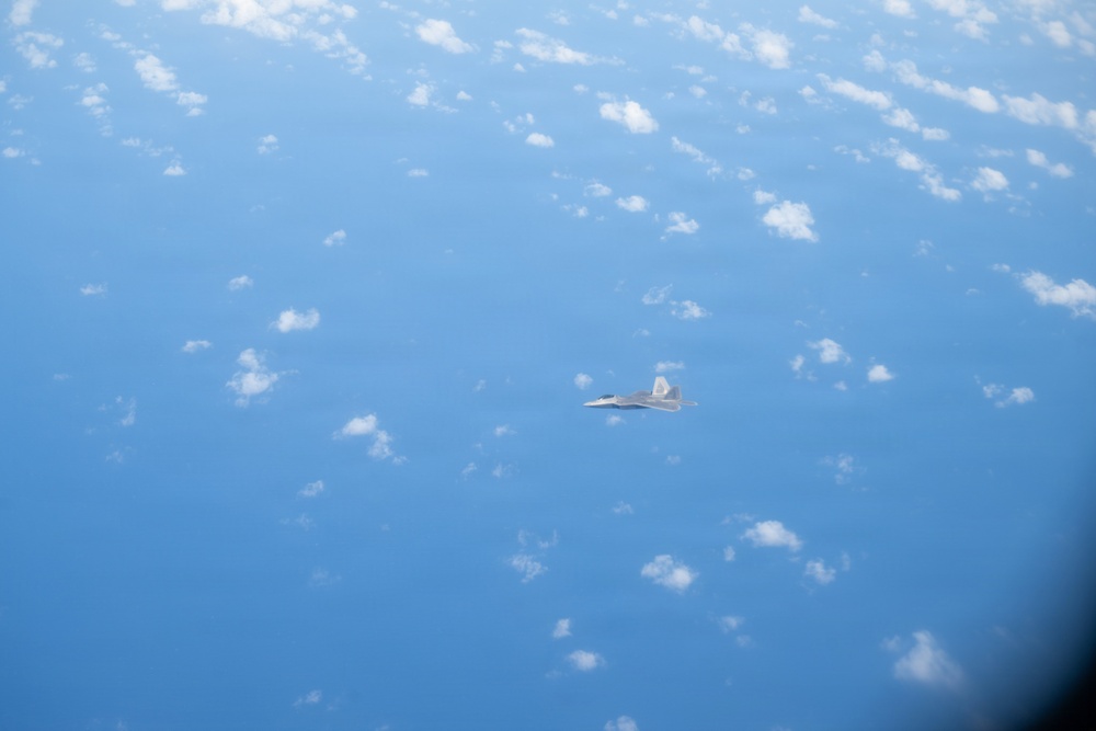 An aerial photograph taken from a high altitude showing a single military jet aircraft flying over a vast expanse of blue ocean or sky with scattered white clouds below. The aircraft appears to be a twin-engine fighter jet, likely an F-22 Raptor based on its distinctive silhouette and delta wing configuration. The image was likely taken from another aircraft flying at a similar or slightly higher altitude.