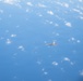 An aerial photograph taken from a high altitude showing a single military jet aircraft flying over a vast expanse of blue ocean or sky with scattered white clouds below. The aircraft appears to be a twin-engine fighter jet, likely an F-22 Raptor based on its distinctive silhouette and delta wing configuration. The image was likely taken from another aircraft flying at a similar or slightly higher altitude.