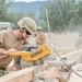 Military personnel in combat uniforms and helmets are engaged in construction work at an outdoor site. The primary subject is operating a DeWalt miter saw to cut lumber, with sawdust visibly dispersing in the air. Multiple service members are working collaboratively in what appears to be a field engineering or humanitarian construction exercise.