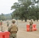 The image shows military personnel in camouflage uniforms manning what appears to be a checkpoint or perimeter security position in a rural, dry landscape. Orange traffic barriers and concertina/razor wire are deployed along a dirt road, suggesting a controlled access point or security exercise. Two individuals in the background appear to be wearing civilian-style hard hats, possibly civilian contractors or observers.