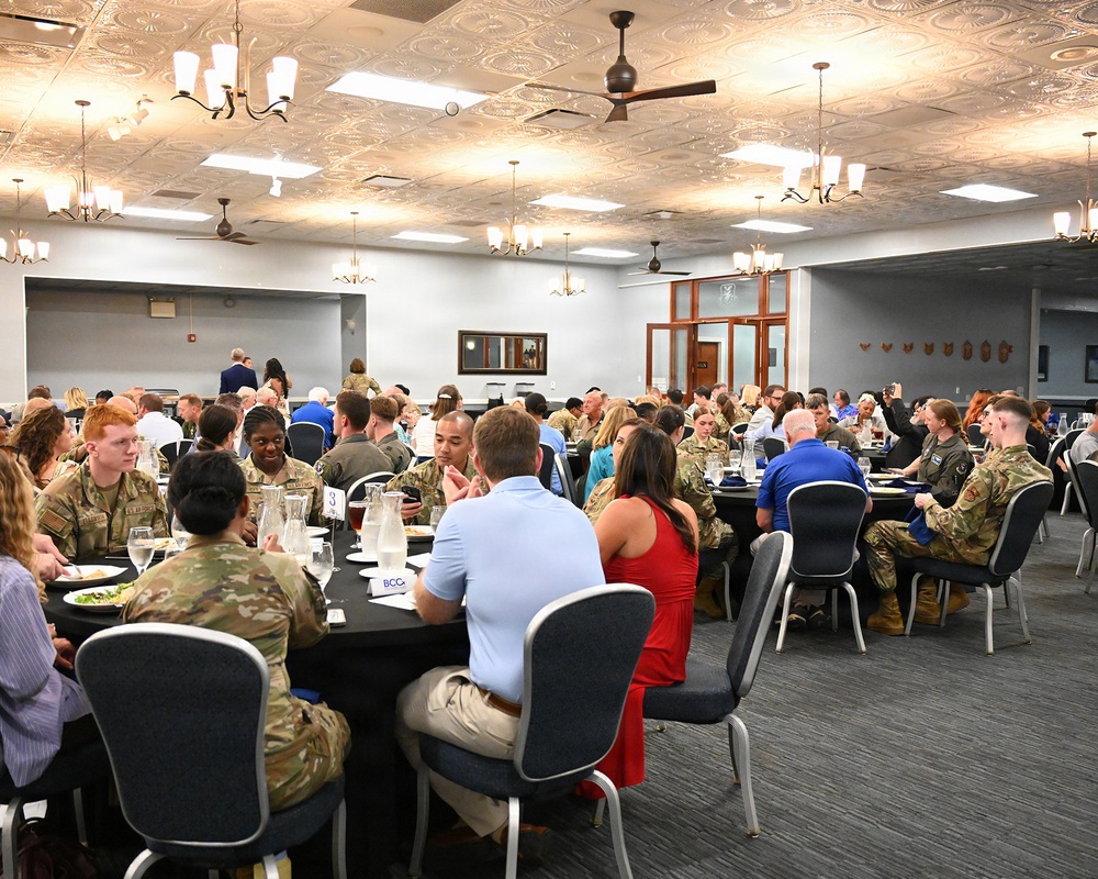 A formal military dining event or luncheon is taking place in a large banquet hall. Numerous military personnel in camouflage uniforms (OCP pattern) are seated at round tables alongside civilians. The setting appears to be an official function, possibly a recognition dinner, community engagement event, or military-civilian relations gathering.