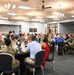 A formal military dining event or luncheon is taking place in a large banquet hall. Numerous military personnel in camouflage uniforms (OCP pattern) are seated at round tables alongside civilians. The setting appears to be an official function, possibly a recognition dinner, community engagement event, or military-civilian relations gathering.