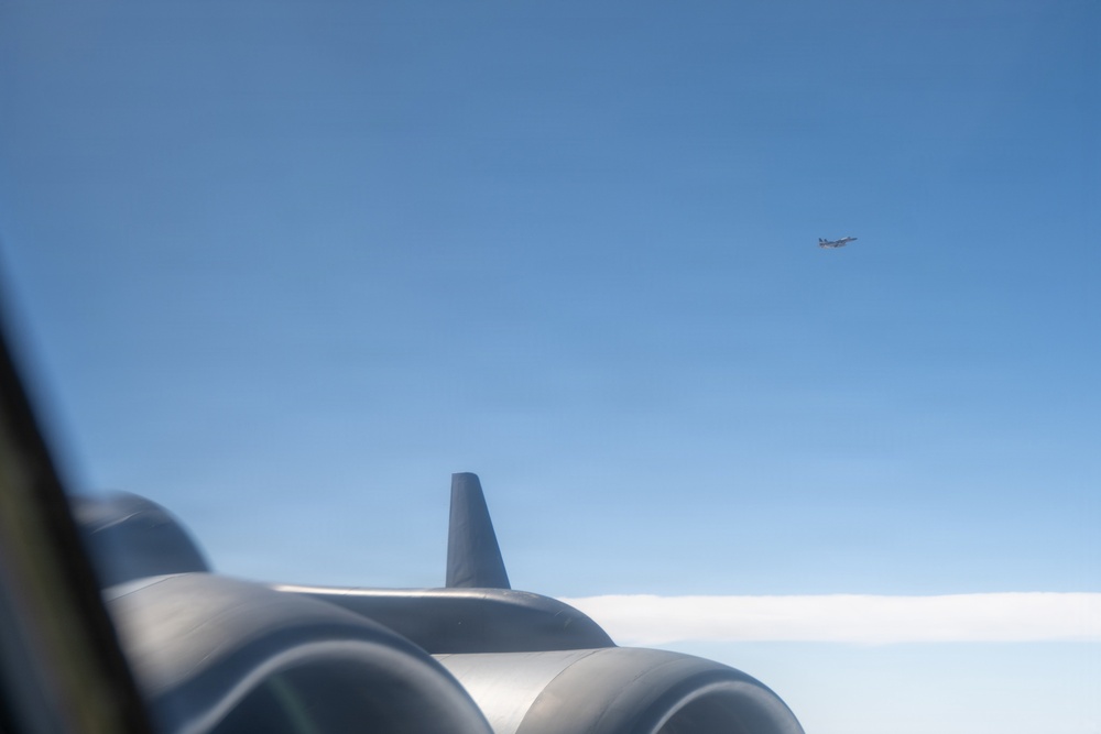 The image is taken from inside or alongside a large multi-engine aircraft, showing its engine nacelles and a small antenna in the foreground. In the background, at least one other aircraft is visible flying at a distance against a clear blue sky. The perspective suggests an aerial formation or escort scenario photographed through or near a window.