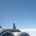 The image is taken from inside or alongside a large multi-engine aircraft, showing its engine nacelles and a small antenna in the foreground. In the background, at least one other aircraft is visible flying at a distance against a clear blue sky. The perspective suggests an aerial formation or escort scenario photographed through or near a window.