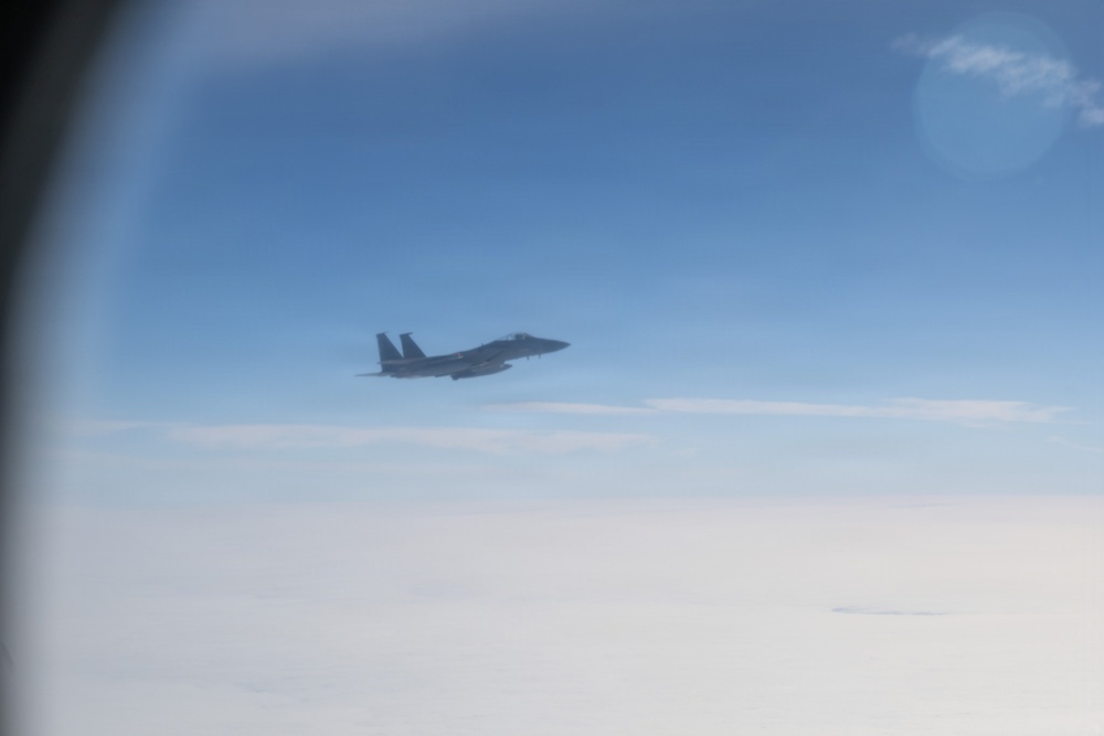 A military jet aircraft, consistent with an F-15 Eagle design, is photographed in flight from what appears to be another aircraft's window. The jet is silhouetted against a blue sky with a cloud layer visible below. The image appears to capture an aerial intercept or escort scenario.