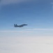 A military jet aircraft, consistent with an F-15 Eagle design, is photographed in flight from what appears to be another aircraft's window. The jet is silhouetted against a blue sky with a cloud layer visible below. The image appears to capture an aerial intercept or escort scenario.