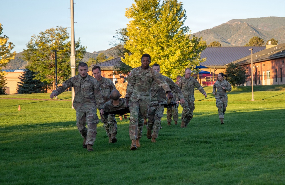 A group of military personnel in OCP (Operational Camouflage Pattern) uniforms are conducting a litter/stretcher carry drill on a grassy field during early morning or evening hours. The exercise appears to be a combat casualty evacuation training exercise, with soldiers carrying a simulated casualty on a litter. The setting appears to be a military installation with brick buildings and mountainous terrain visible in the background.