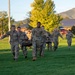 A group of military personnel in OCP (Operational Camouflage Pattern) uniforms are conducting a litter/stretcher carry drill on a grassy field during early morning or evening hours. The exercise appears to be a combat casualty evacuation training exercise, with soldiers carrying a simulated casualty on a litter. The setting appears to be a military installation with brick buildings and mountainous terrain visible in the background.