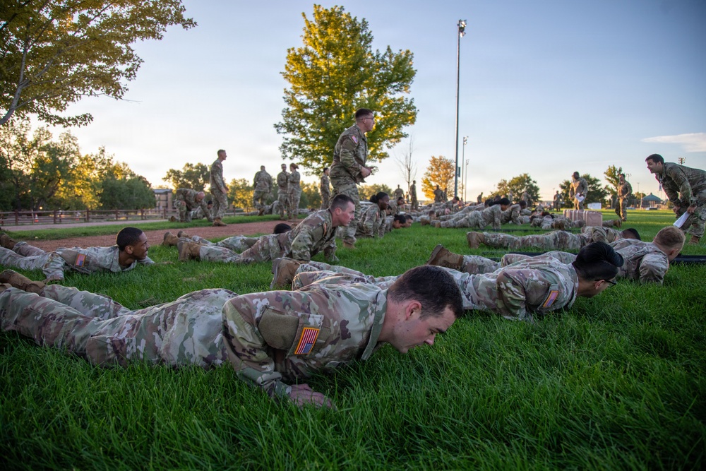 A large group of U.S. Army soldiers in OCP (Operational Camouflage Pattern) uniforms are performing push-ups on a grass field during what appears to be an early morning physical fitness training session. Several supervisory personnel are standing and walking among the rows of soldiers doing push-ups, likely counting repetitions or monitoring form. The setting appears to be a military installation training field at dawn or dusk.