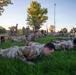 A large group of U.S. Army soldiers in OCP (Operational Camouflage Pattern) uniforms are performing push-ups on a grass field during what appears to be an early morning physical fitness training session. Several supervisory personnel are standing and walking among the rows of soldiers doing push-ups, likely counting repetitions or monitoring form. The setting appears to be a military installation training field at dawn or dusk.