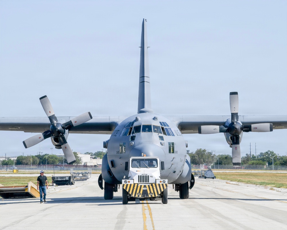 A large military transport aircraft, identified as a U.S. Air Force C-130 Hercules, is being towed along a taxiway by a yellow-and-black striped aircraft tug vehicle. The aircraft is viewed head-on, displaying its four turboprop engines with large propeller blades. A ground crew member in civilian attire walks alongside the aircraft on the left side.