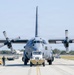 A large military transport aircraft, identified as a U.S. Air Force C-130 Hercules, is being towed along a taxiway by a yellow-and-black striped aircraft tug vehicle. The aircraft is viewed head-on, displaying its four turboprop engines with large propeller blades. A ground crew member in civilian attire walks alongside the aircraft on the left side.