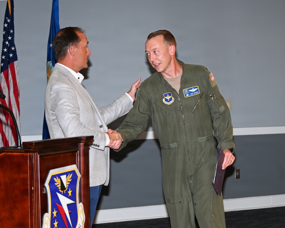 A formal military ceremony is taking place at what appears to be a U.S. Air Force installation. A civilian in a gray blazer is shaking hands with and placing a hand on the shoulder of a military aviator in an olive green flight suit. The aviator is holding what appears to be a certificate or folder, suggesting an award or recognition ceremony.