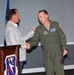 A formal military ceremony is taking place at what appears to be a U.S. Air Force installation. A civilian in a gray blazer is shaking hands with and placing a hand on the shoulder of a military aviator in an olive green flight suit. The aviator is holding what appears to be a certificate or folder, suggesting an award or recognition ceremony.