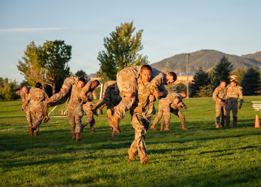Military personnel in camouflage uniforms are conducting physical training exercises on a grass field, with multiple pairs performing buddy carries and combat fitness drills. The scene appears to be an outdoor PT session during golden hour lighting, set against a backdrop of trees, mountains, and a clear sky. The training involves soldiers carrying fellow soldiers on their backs in a coordinated group exercise.