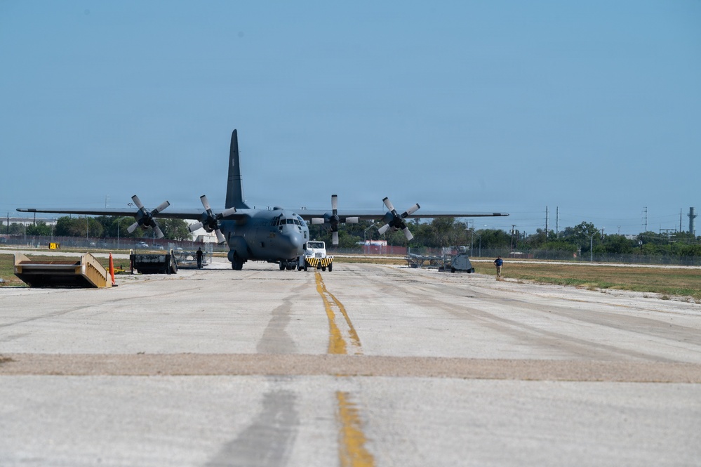 A military C-130 Hercules turboprop aircraft is positioned on a taxiway or apron at a military airfield. A yellow-and-black tow vehicle is visible behind the aircraft, suggesting it is being towed or repositioned. Ground personnel and support equipment are visible on both sides of the aircraft.