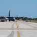 A military C-130 Hercules turboprop aircraft is positioned on a taxiway or apron at a military airfield. A yellow-and-black tow vehicle is visible behind the aircraft, suggesting it is being towed or repositioned. Ground personnel and support equipment are visible on both sides of the aircraft.