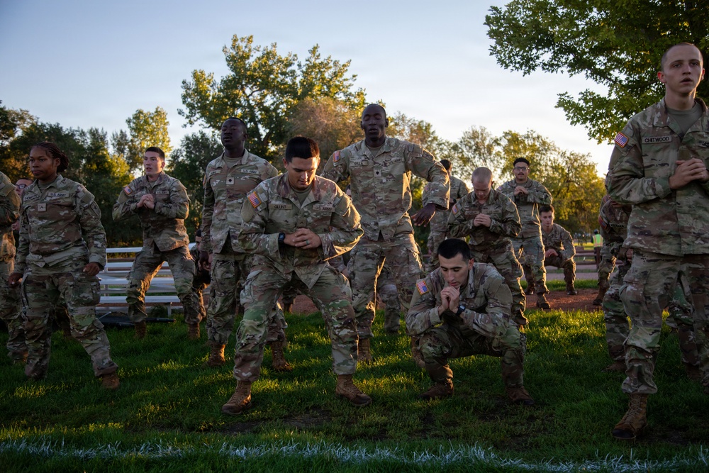 A group of U.S. Army soldiers in OCP (Operational Camouflage Pattern) uniforms are conducting physical training exercises outdoors on a grass field during early morning or evening hours. The soldiers are performing what appears to be a synchronized squat or lower-body exercise as part of a group PT (Physical Training) session. The warm golden light suggests dawn or dusk conditions.
