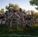A group of U.S. Army soldiers in OCP (Operational Camouflage Pattern) uniforms are conducting physical training exercises outdoors on a grass field during early morning or evening hours. The soldiers are performing what appears to be a synchronized squat or lower-body exercise as part of a group PT (Physical Training) session. The warm golden light suggests dawn or dusk conditions.