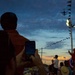 A crowd of spectators at an outdoor event, likely an airshow, watches and photographs a large military aircraft flying overhead at dusk. The scene is captured in low-light conditions with a dramatic blue and orange sunset sky. Multiple people are using smartphones and devices to document the flyover.