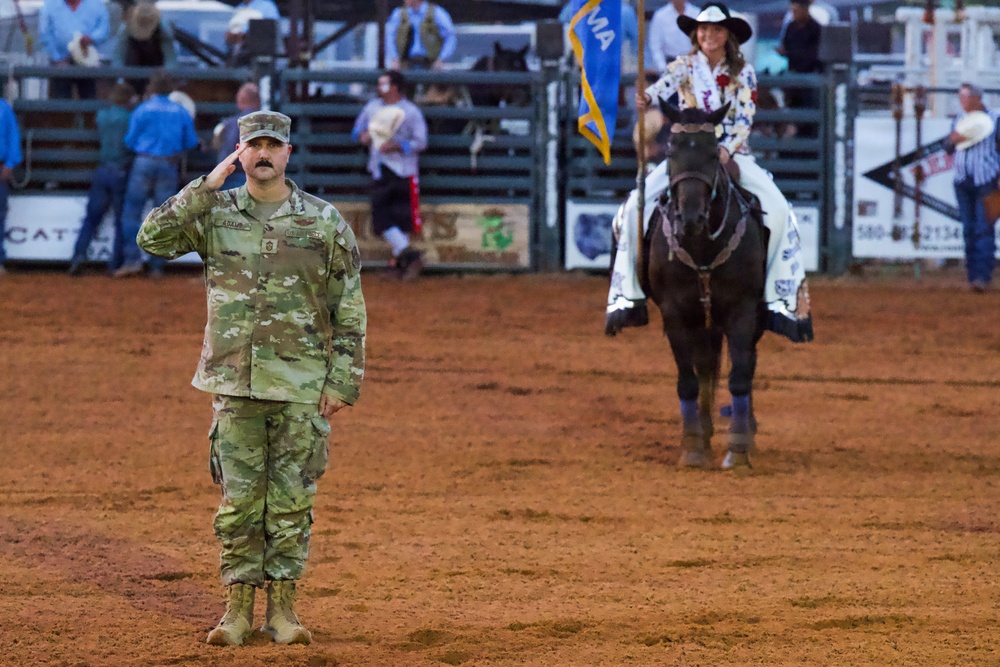 A U.S. Army soldier in MultiCam (OCP) camouflage uniform stands at attention rendering a salute in a rodeo arena. Behind him, a woman on a dark horse carries what appears to be a state flag, suggesting this is an opening ceremony or color presentation. The setting is an indoor/outdoor rodeo venue with spectators visible in the background.