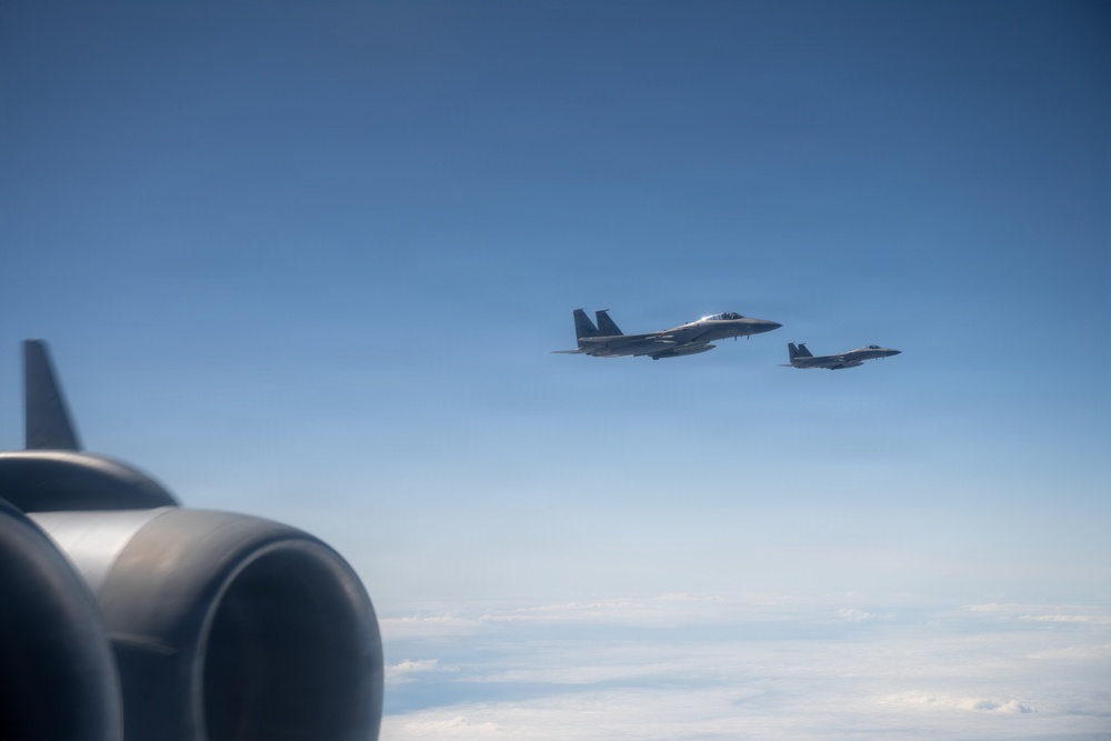 Two military fighter jets, identifiable as F-15 Eagles, are flying in formation against a clear blue sky. The photograph is taken from aboard another aircraft, with a jet engine nacelle and vertical stabilizer visible in the foreground. The aircraft appear to be on a escort or formation flight mission at high altitude above a cloud layer.