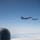 Two military fighter jets, identifiable as F-15 Eagles, are flying in formation against a clear blue sky. The photograph is taken from aboard another aircraft, with a jet engine nacelle and vertical stabilizer visible in the foreground. The aircraft appear to be on a escort or formation flight mission at high altitude above a cloud layer.