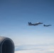 Two military fighter jets, identifiable as F-15 Eagles, are flying in formation against a clear blue sky. The photograph is taken from aboard another aircraft, with a jet engine nacelle and vertical stabilizer visible in the foreground. The aircraft appear to be on a escort or formation flight mission at high altitude above a cloud layer.