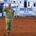 A U.S. Air Force military officer in OCP (Operational Camouflage Pattern) uniform stands at attention and salutes in the middle of a rodeo arena with red dirt ground. The name tape on the uniform appears to read 'MOORES' and rank insignia is visible on the chest. The setting is an outdoor rodeo event in Altus, Oklahoma, as indicated by sponsor banners in the background.