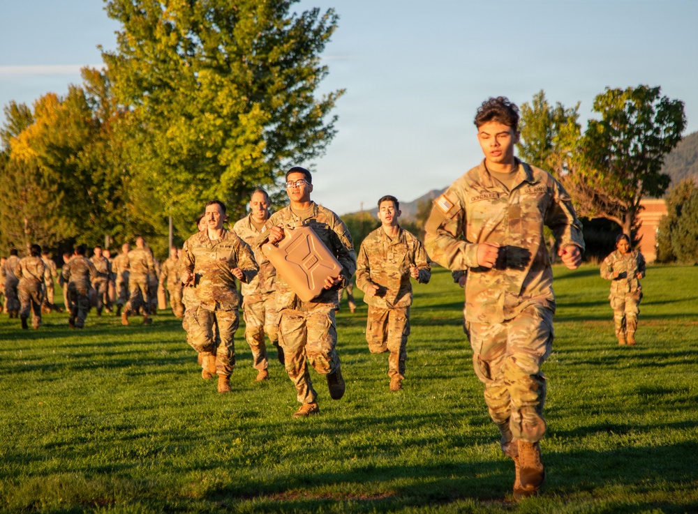 A group of U.S. Army soldiers in OCP (Operational Camouflage Pattern) uniforms are running across a grassy field during what appears to be a physical training exercise at golden hour. The lead soldier, whose name tape reads 'GONZALEZ,' is running in front of a large formation. One soldier in the middle group is carrying a tan military-style jerry can as part of the exercise.