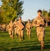 A group of U.S. Army soldiers in OCP (Operational Camouflage Pattern) uniforms are running across a grassy field during what appears to be a physical training exercise at golden hour. The lead soldier, whose name tape reads 'GONZALEZ,' is running in front of a large formation. One soldier in the middle group is carrying a tan military-style jerry can as part of the exercise.
