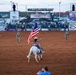 A rodeo opening ceremony is taking place at the Great Plains Stampede Rodeo in Altus, Oklahoma. A rider on a white horse carries the American flag across the dirt arena while military personnel in camouflage uniforms stand at attention around the perimeter. The event features multiple sponsor banners and a grandstand with spectators.