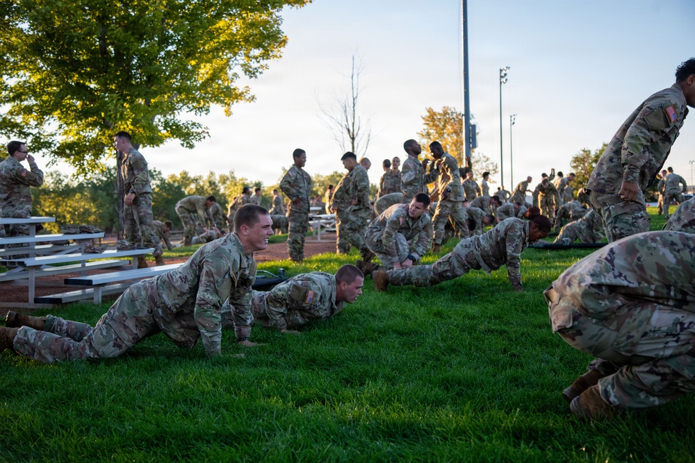 A large group of military personnel in OCP (Operational Camouflage Pattern) uniforms are conducting physical training on a grass field. Several soldiers in the foreground are performing push-ups while others stand or engage in various exercises in the background. The setting appears to be an outdoor athletic or parade field during early morning or evening hours based on the lighting.