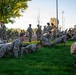 A large group of military personnel in OCP (Operational Camouflage Pattern) uniforms are conducting physical training on a grass field. Several soldiers in the foreground are performing push-ups while others stand or engage in various exercises in the background. The setting appears to be an outdoor athletic or parade field during early morning or evening hours based on the lighting.